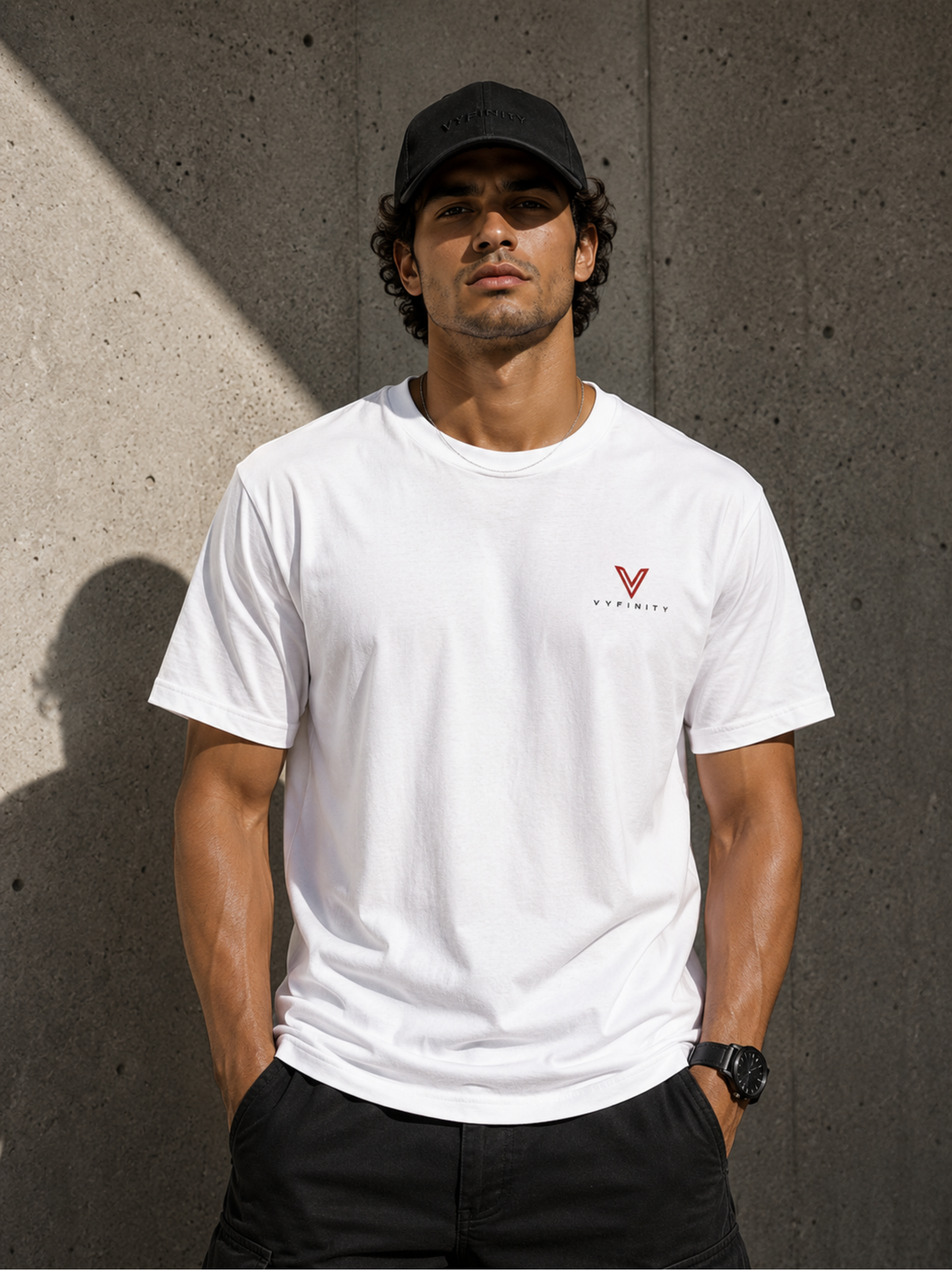 Männliches Model mit schwarzer VYFINITY Cap und weißem VYFINITY T-Shirt mit kleinem rot-schwarzem Brustlogo, vor moderner Betonwand im Sonnenlicht.