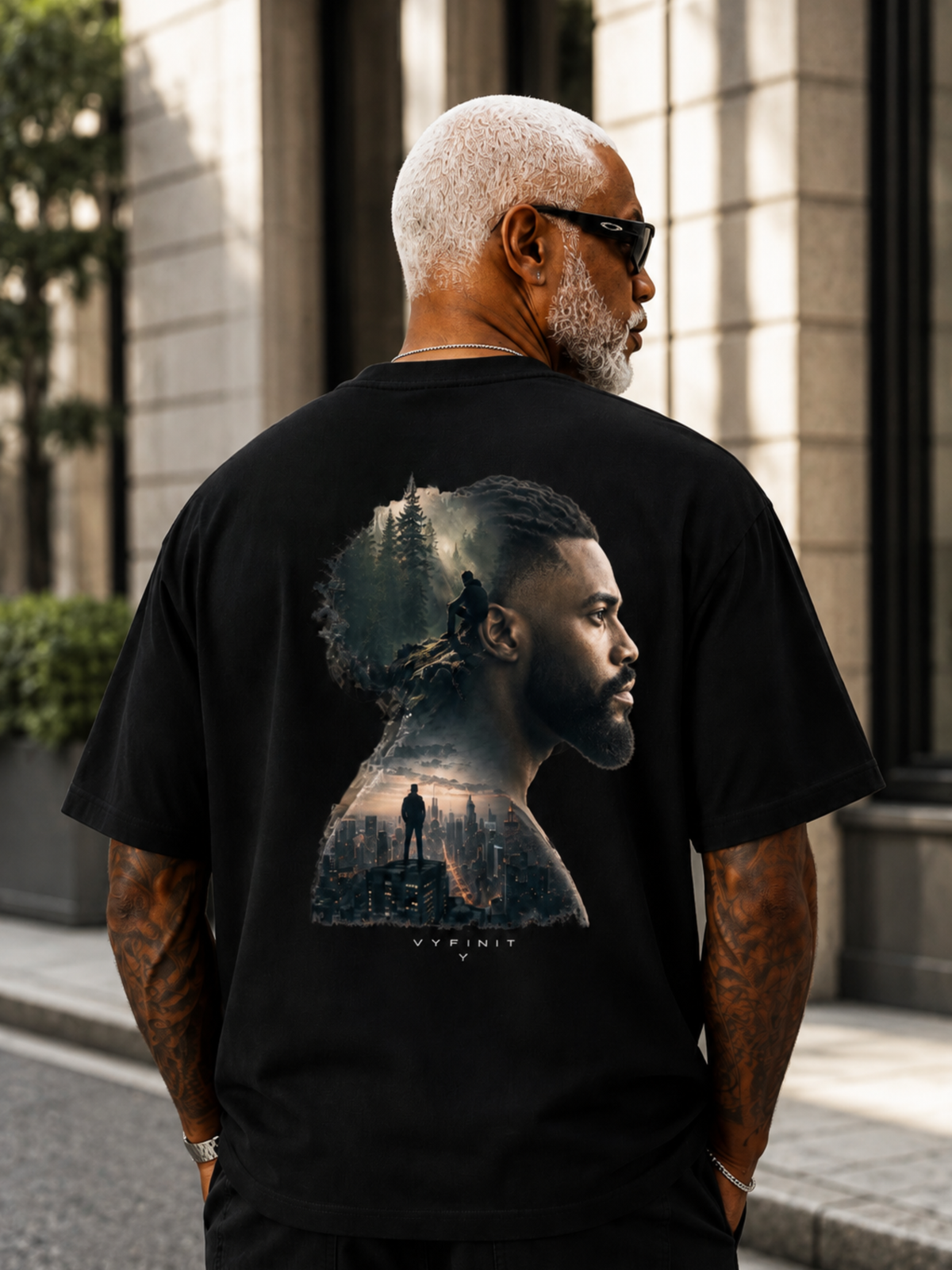 Älterer männlicher Model trägt ein schwarzes VYFINITY Heavy T-Shirt in Rückansicht. Großer Double-Exposure-Rückenprint mit männlichem Seitenprofil, Waldlandschaft, einsamer Figur und nächtlicher Skyline. Urbanes Lifestyle-Shooting vor moderner Architektur.