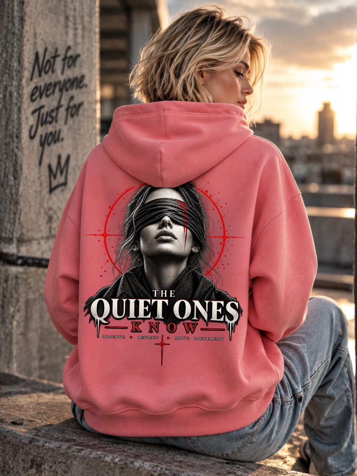 Blondes weibliches Model trägt einen pinken Oversized Hoodie mit großem THE QUIET ONES KNOW Rückenprint, Frauenporträt mit schwarzer Augenbinde und roten grafischen Elementen, in urbaner Kulisse bei Sonnenuntergang.