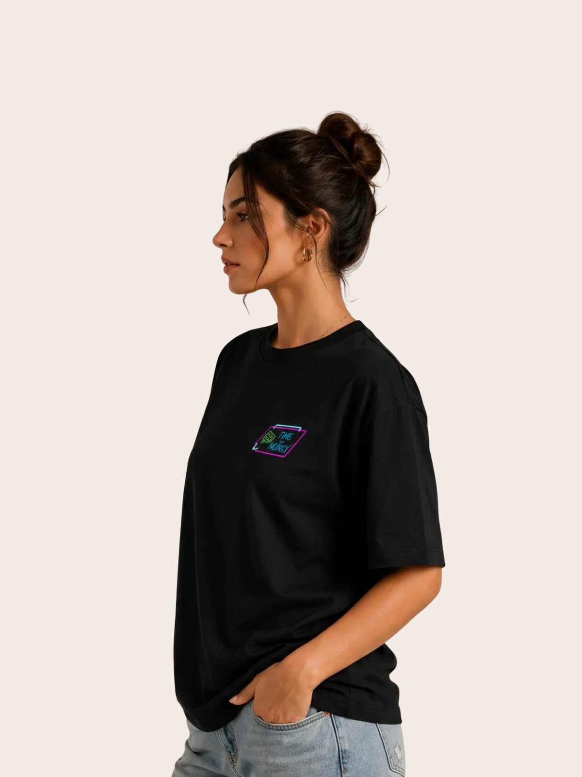 Frau im Seitenprofil trägt ein schwarzes VYFINITY T-Shirt mit kleinem neonfarbenem „TIME MONEY“-Brustprint in Pink, Blau und Grün. Lockerer Streetwear-Fit mit heller Jeans vor neutralem Hintergrund.