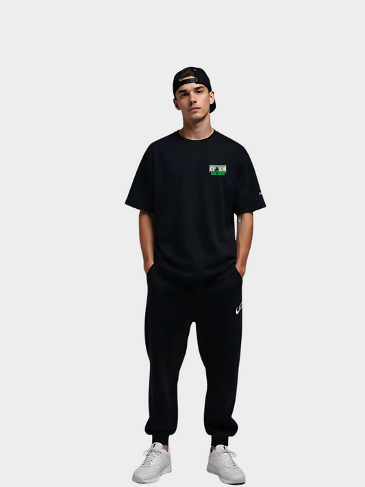 Frontansicht eines schwarzen Vyfinity T-Shirts „Cash Coded“ mit kleinem grünen Brustlogo, getragen von einem Model mit schwarzer Cap, schwarzer Hose und weißen Sneakern.
