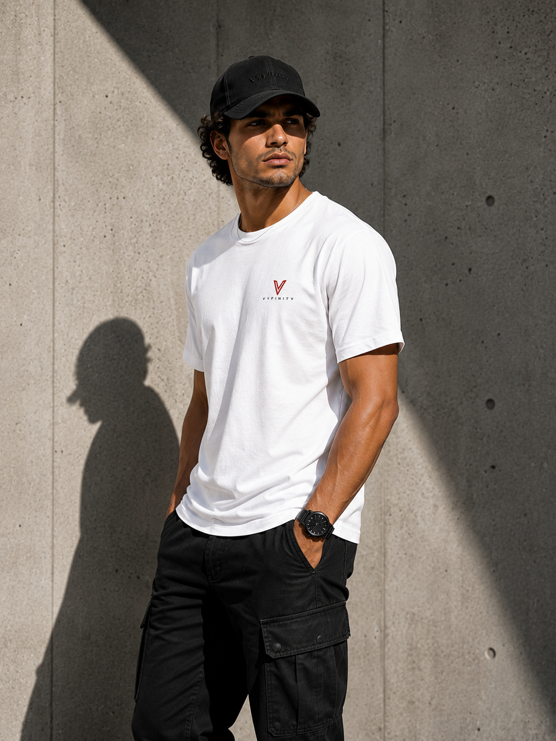 Männliches Model mit schwarzer VYFINITY Cap und weißem VYFINITY T-Shirt mit kleinem rot-schwarzem Brustlogo, seitlich vor moderner Betonwand im direkten Sonnenlicht. Schwarze Cargo-Hose und Uhr ergänzen den minimalistischen Streetwear-Look.