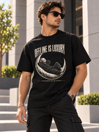 Schwarzes Oversized Heavy T-Shirt mit großem „OFFLINE IS LUXURY“-Frontprint. Zentral liegt eine entspannte Alien-Figur im schwarzen Hoodie mit Kopfhörern auf einer realistisch strukturierten Mondsichel. Darunter „DISCONNECTED BY CHOICE“. Moderner monochromer Streetwear-Look in Creme, Grau und Schwarz mit dezenten warmen Akzenten.