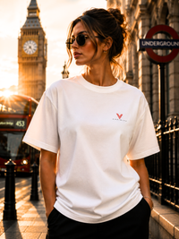 Frau trägt ein weißes VYFINITY Oversized Unisex T-Shirt mit rotem Brustlogo vor dem Big Ben und einer London Underground Station bei Sonnenuntergang. Modernes Streetwear Lifestyle-Foto mit rotem Doppeldeckerbus, urbanem City-Flair und minimalistischem Premium Fashion Look.