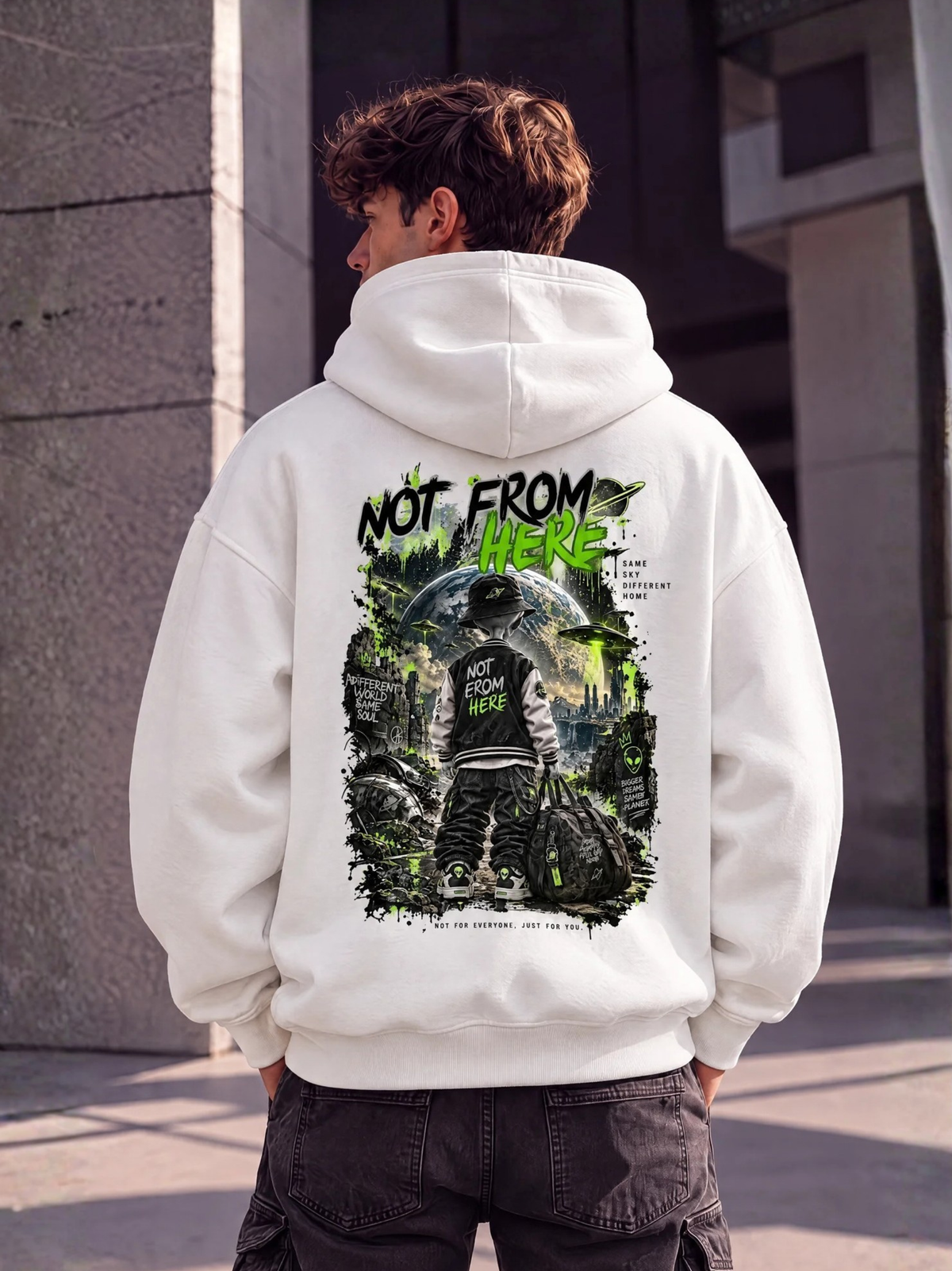 Männliches Model trägt den weißen VYFINITY Alien Not From Here Hoodie mit großem schwarz-grünem Alien-Rückenprint und dem Schriftzug NOT FROM HERE vor moderner urbaner Architektur.