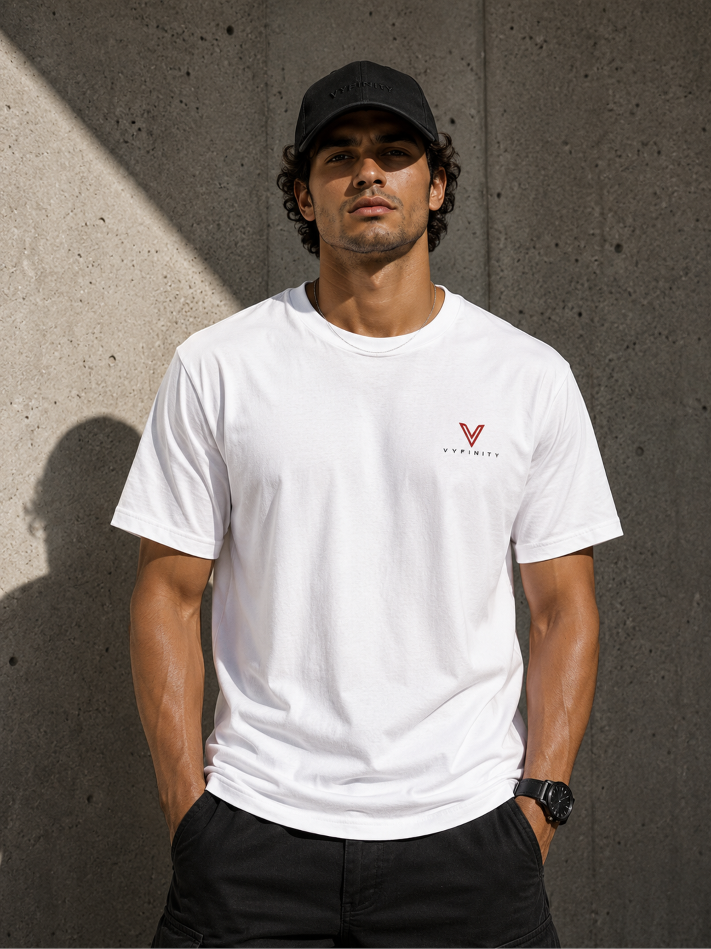Männliches Model mit schwarzer VYFINITY Cap und weißem VYFINITY T-Shirt mit kleinem rot-schwarzem Brustlogo, vor moderner Betonwand im Sonnenlicht.