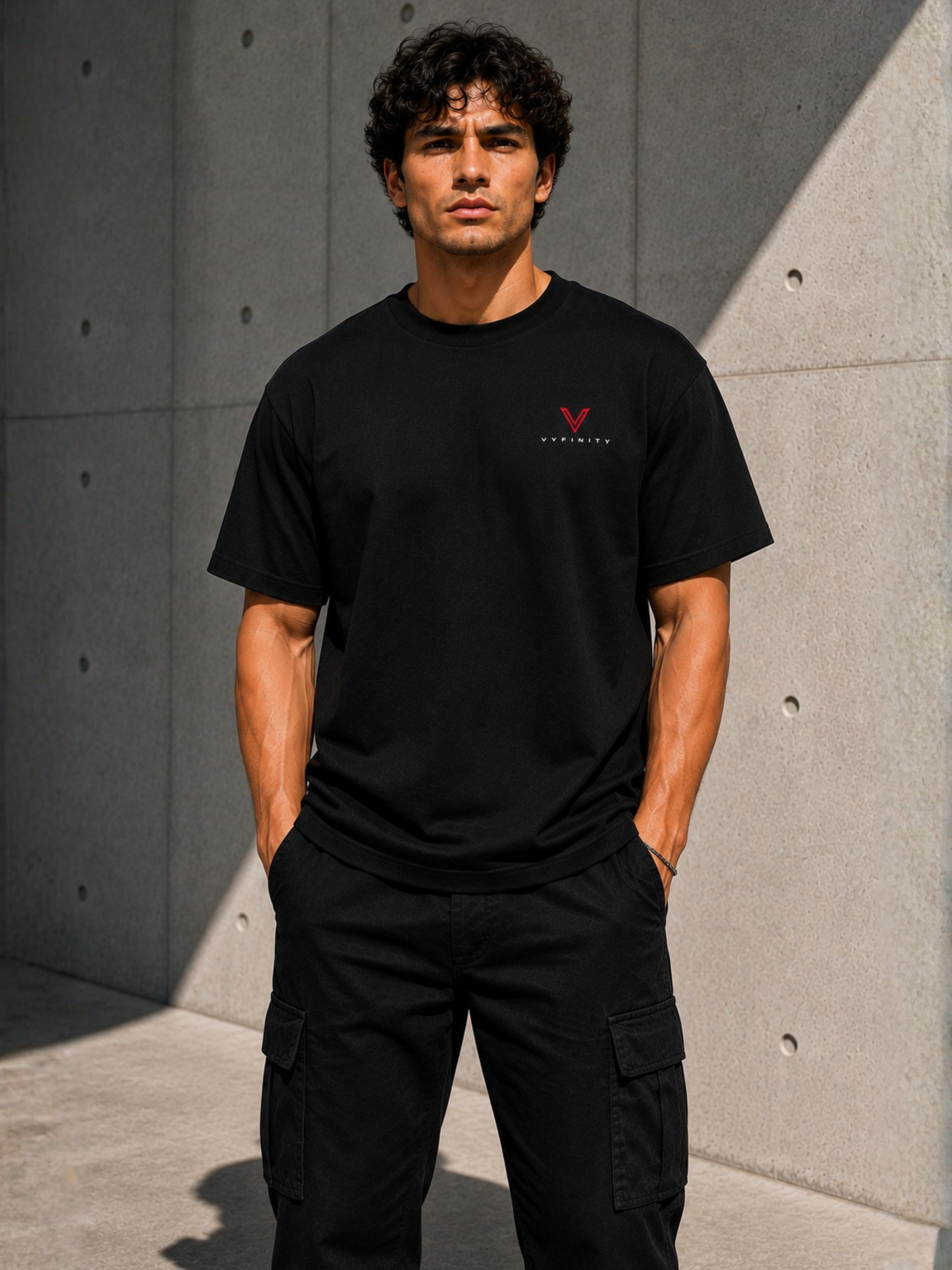 Männliches Model trägt ein schwarzes VYFINITY T-Shirt mit kleinem rot-weißem Brustlogo und schwarze Cargo-Hose vor moderner Betonwand.