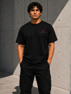 Männliches Model trägt ein schwarzes VYFINITY T-Shirt mit kleinem rot-weißem Brustlogo und schwarze Cargo-Hose vor moderner Betonwand.