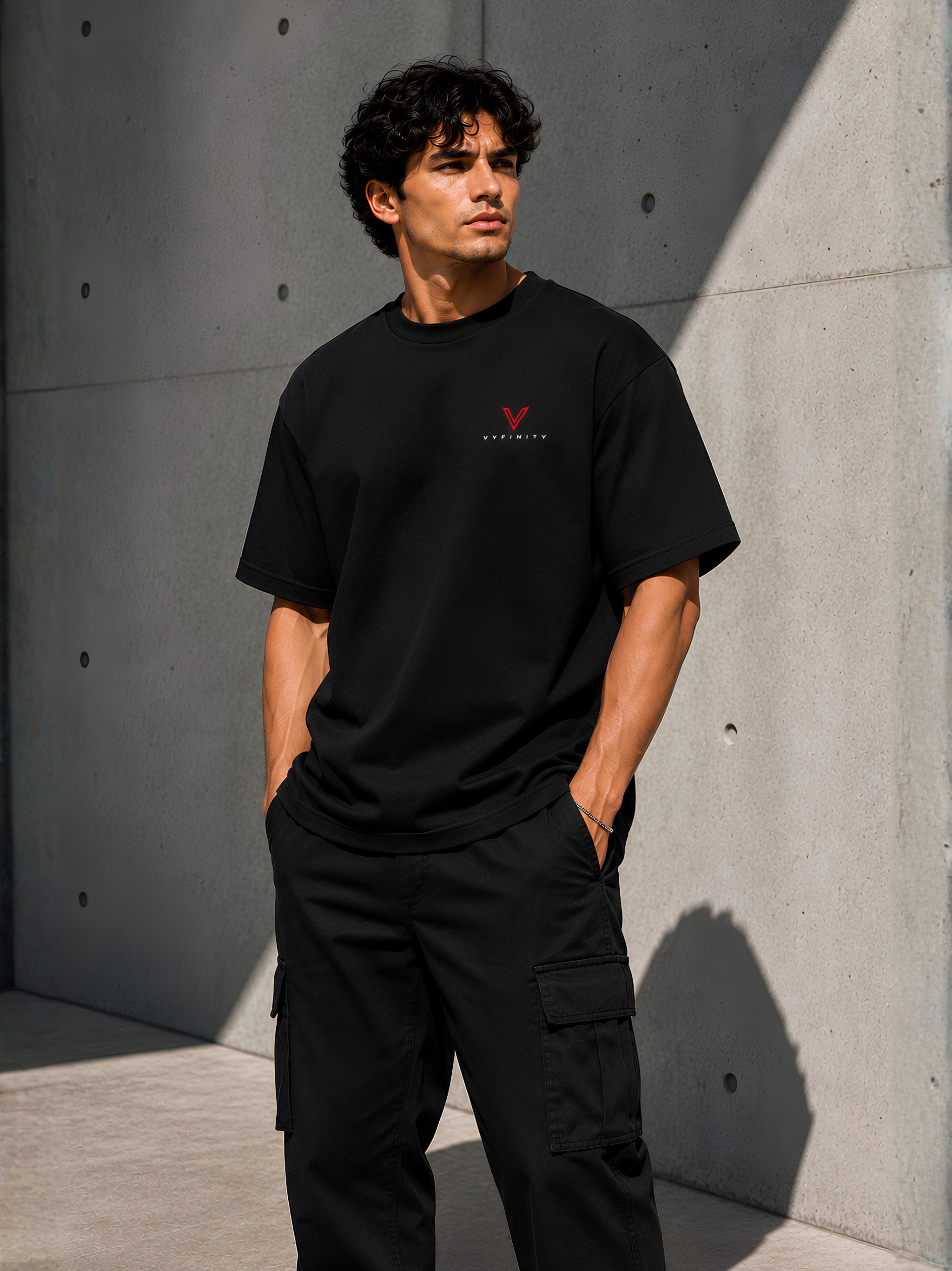 Männliches Model in schwarzem VYFINITY T-Shirt mit kleinem rot-weißem Brustlogo und schwarzer Cargo-Hose, seitlich vor moderner Betonwand im Sonnenlicht.