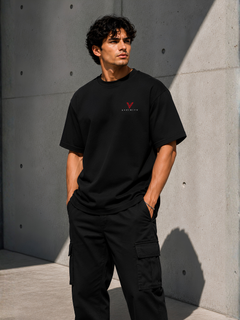 Männliches Model in schwarzem VYFINITY T-Shirt mit kleinem rot-weißem Brustlogo und schwarzer Cargo-Hose, seitlich vor moderner Betonwand im Sonnenlicht.