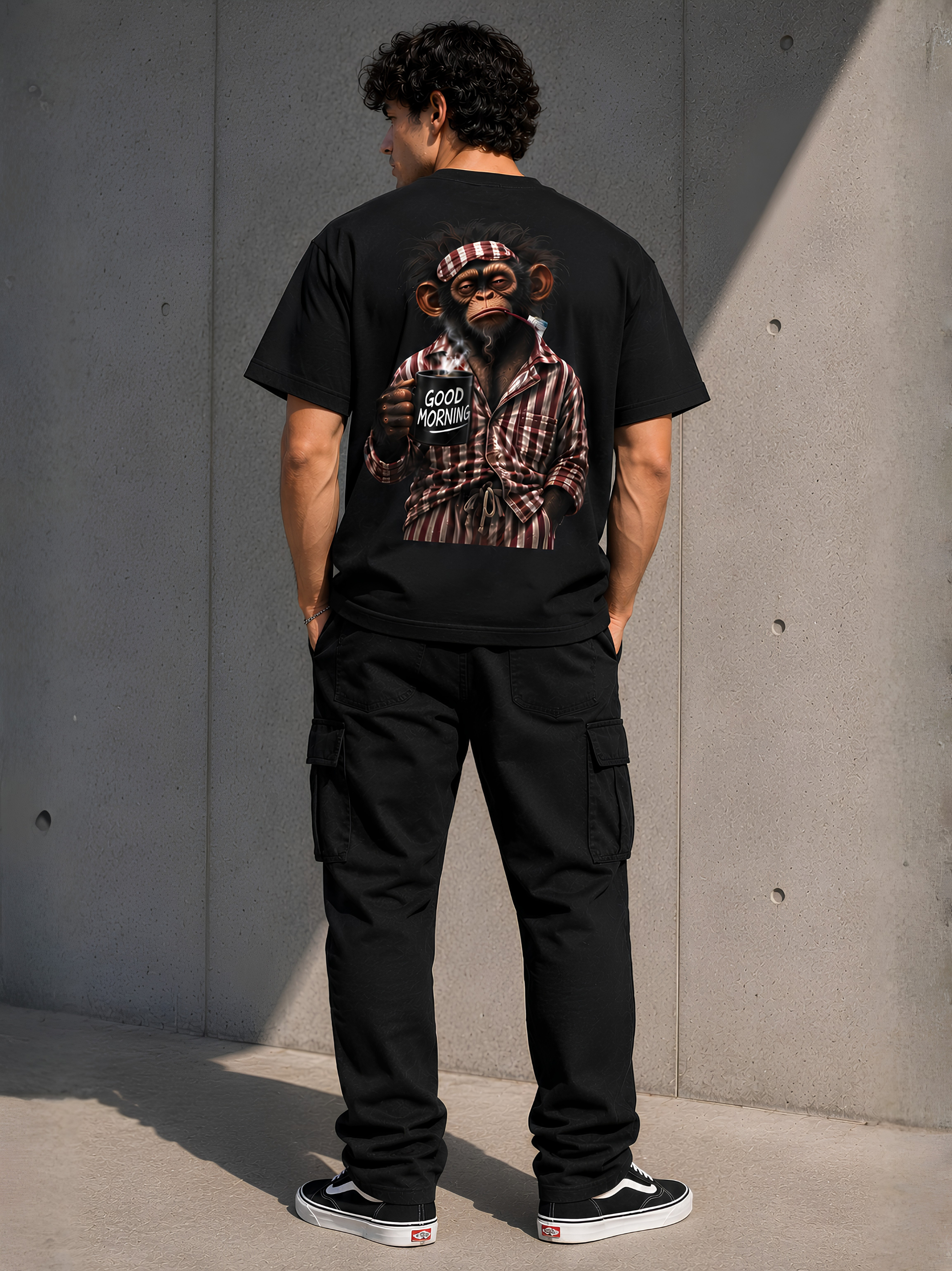 Männliches Model in schwarzem VYFINITY T-Shirt in Rückansicht mit großem „Good Morning“-Affenmotiv, schwarzer Cargo-Hose und Sneakern vor moderner Betonwand.