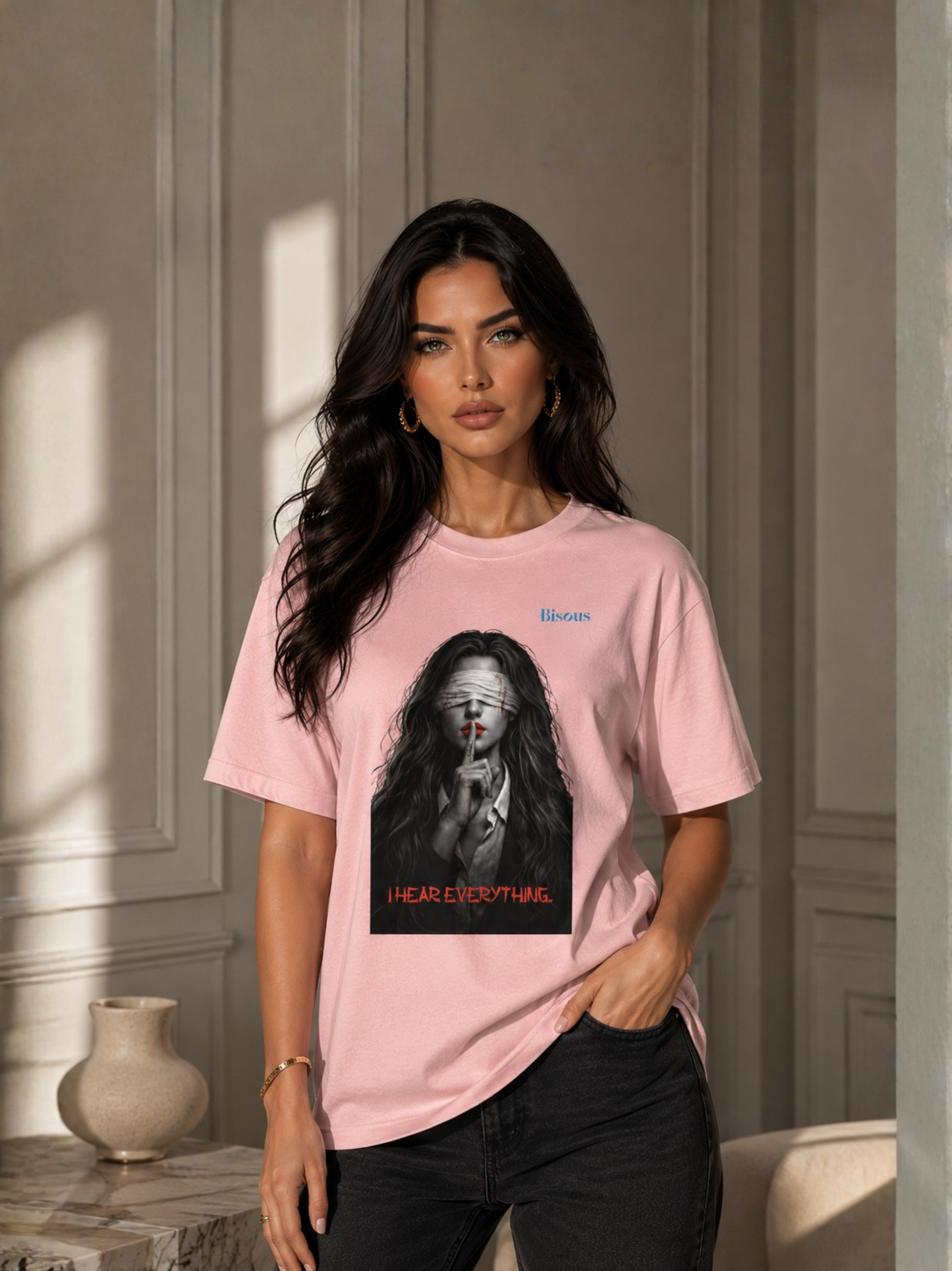 Elegante Frau trägt ein rosa Unisex Classic Fit T-Shirt mit markantem Dark-Art-Motiv einer verbundenen Frau mit roten Lippen, Finger vor dem Mund und dem Schriftzug „I Hear Everything“. Modernes Premium Baumwoll-T-Shirt im Gothic-, Horror- und Streetwear-Stil, fotografiert in einem stilvollen Interior mit natürlichem Tageslicht.