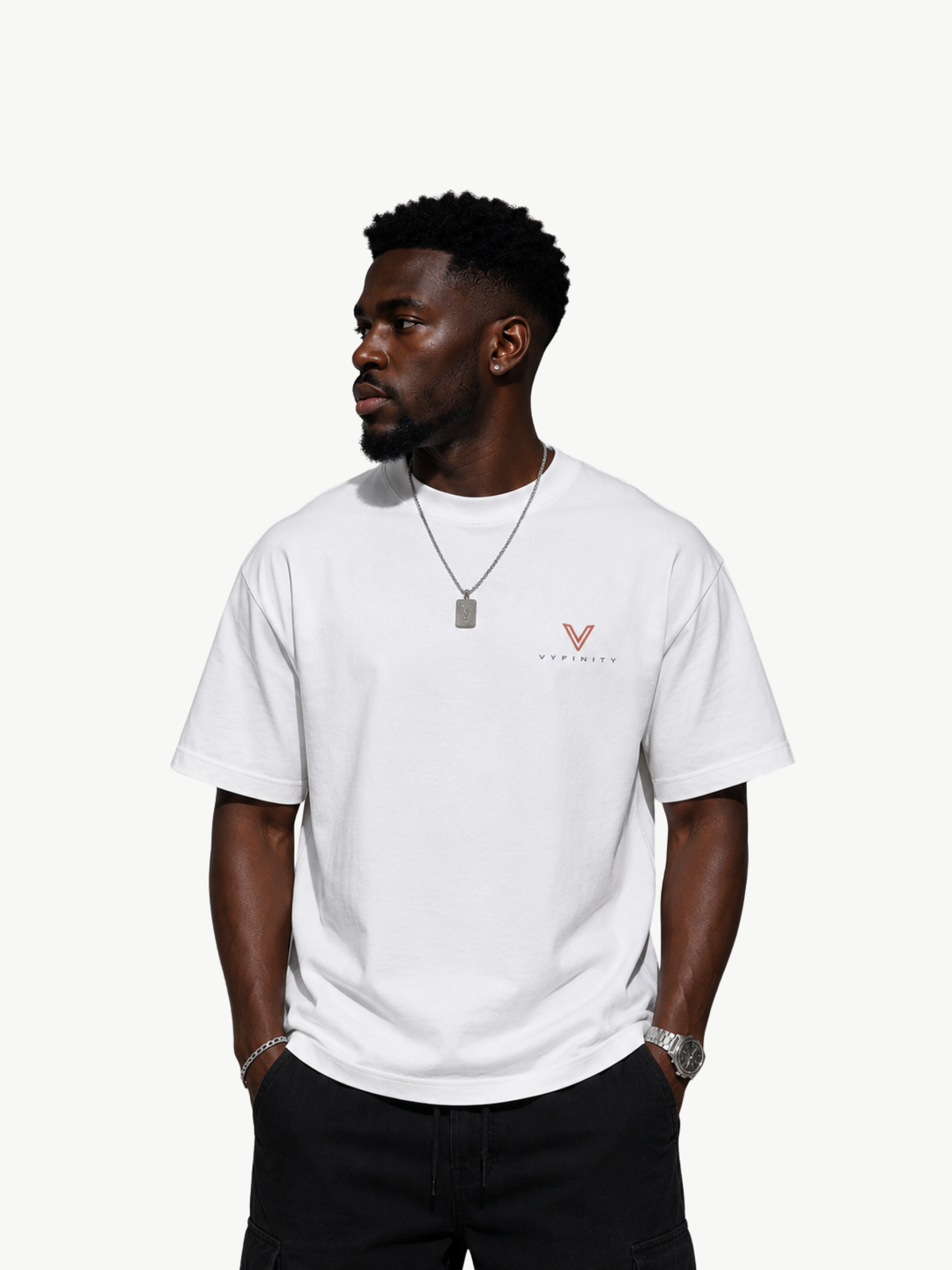 Männliches Model trägt ein weißes VYFINITY Heavy T-Shirt in Frontansicht mit kleinem rot-weißem VYFINITY-Logo auf der linken Brust. Minimalistischer Streetwear-Look mit schwarzer Hose und silbernen Accessoires vor hellem Studiohintergrund.