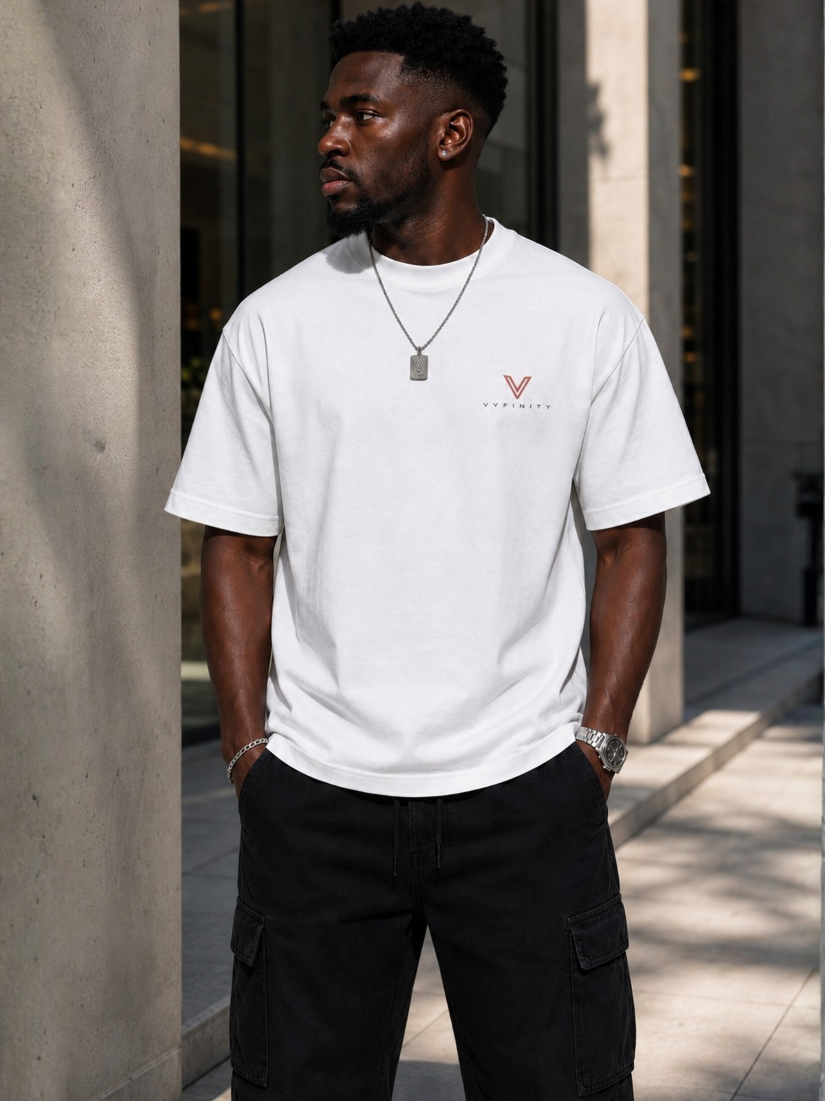 Schwarzer Mann trägt ein weißes VYFINITY Heavy T-Shirt mit kleinem VYFINITY-Logo auf der linken Brust. Urbanes Lifestyle-Foto vor moderner Architektur, kombiniert mit schwarzer Cargo-Hose, silberner Kette und Uhr. Cleanes Premium-Streetwear-Outfit in natürlichem Tageslicht.