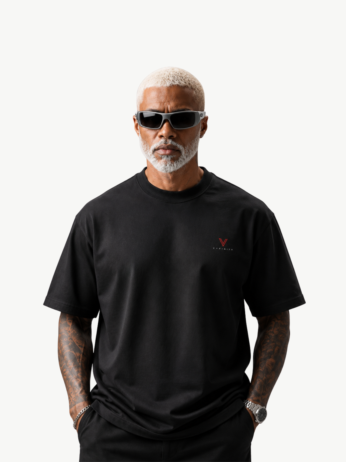  Männliches Model mit blondiertem Kurzhaar, weißem Bart und schwarzer Sonnenbrille trägt ein schwarzes VYFINITY Heavy T-Shirt mit kleinem rot-schwarzem VYFINITY-Logo auf der Brust. Minimalistische Studioaufnahme vor hellem Hintergrund.