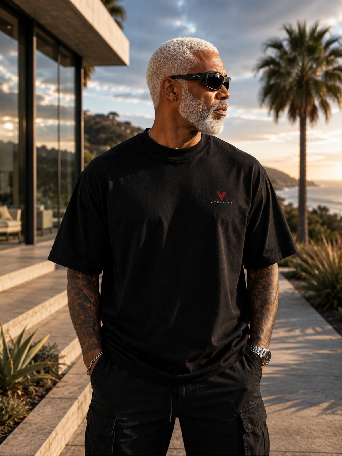 Älterer männlicher Model trägt ein schwarzes VYFINITY Heavy T-Shirt mit kleinem rot-weißem Brustlogo. Lifestyle-Aufnahme vor moderner Villa mit Palmen und Küstenlandschaft im warmen Abendlicht.