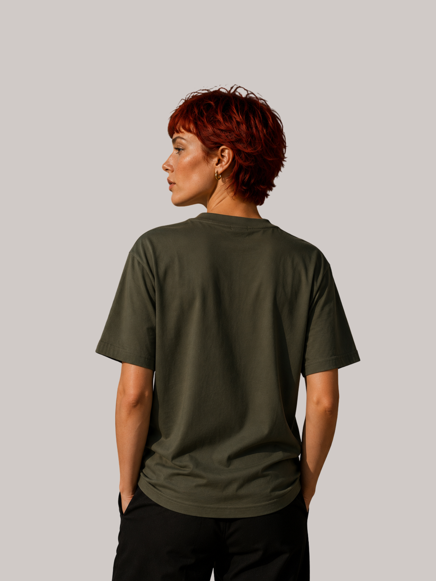 Rückansicht einer jungen Frau mit kurzen roten Haaren im olivgrünen VYFINITY Unisex T-Shirt. Das Shirt zeigt die schlichte Rückseite ohne Frontprint und präsentiert den lockeren Regular Fit in einer modernen Streetwear-Ansicht vor neutralem Studiohintergrund.