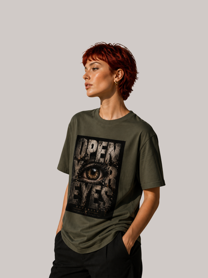 Junge Frau mit kurzen roten Haaren trägt ein olivgrünes VYFINITY Unisex T-Shirt mit großem „OPEN YOUR EYES“-Print und realistischem Auge im markanten Grunge-Design. Seitliche Ansicht im modernen Streetwear-Look vor neutralem Studiohintergrund.