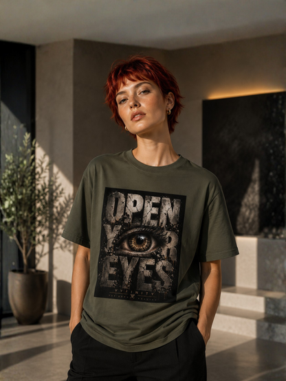 Junge Frau mit kurzen roten Haaren trägt ein olivgrünes VYFINITY Regular Fit T-Shirt mit großem „OPEN YOUR EYES“-Print und realistischem Auge auf der Vorderseite. Das moderne Streetwear-Shirt wird in einem hochwertigen Lifestyle-Setting mit luxuriöser Architektur und warmem Sonnenlicht präsentiert.