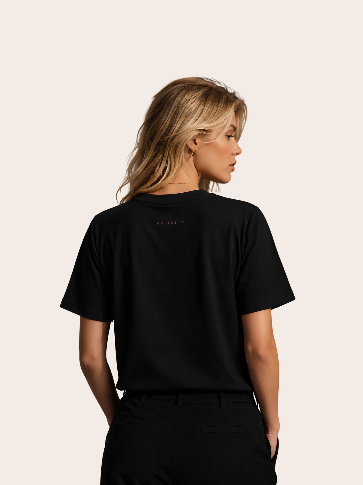 Blonde Frau in Rückansicht trägt ein schwarzes VYFINITY T-Shirt mit kleinem, dezent goldfarbenem VYFINITY-Schriftzug im oberen Rückenbereich. Elegantes schwarzes Ton-in-Ton-Outfit vor hellem, neutralem Hintergrund.