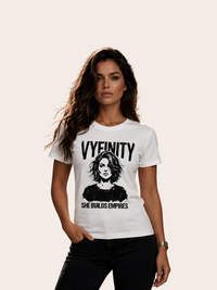 Frau mit langen dunklen Haaren trägt ein weißes VYFINITY T-Shirt mit markantem schwarz-weißem Frauenporträt, großem VYFINITY-Schriftzug und dem Statement „SHE BUILDS EMPIRES“. Moderner monochromer Fashion-Look mit schwarzer Hose vor hellem, neutralem Hintergrund.