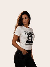 Frau mit langen dunklen Haaren trägt ein weißes VYFINITY T-Shirt mit schwarzem Graphic-Print eines stilisierten Frauenporträts, großem VYFINITY-Schriftzug und dem Statement „SHE BUILDS EMPIRES“. Kombiniert mit schwarzer Jeans in einer seitlichen Fashion-Pose vor hellem, neutralem Hintergrund.