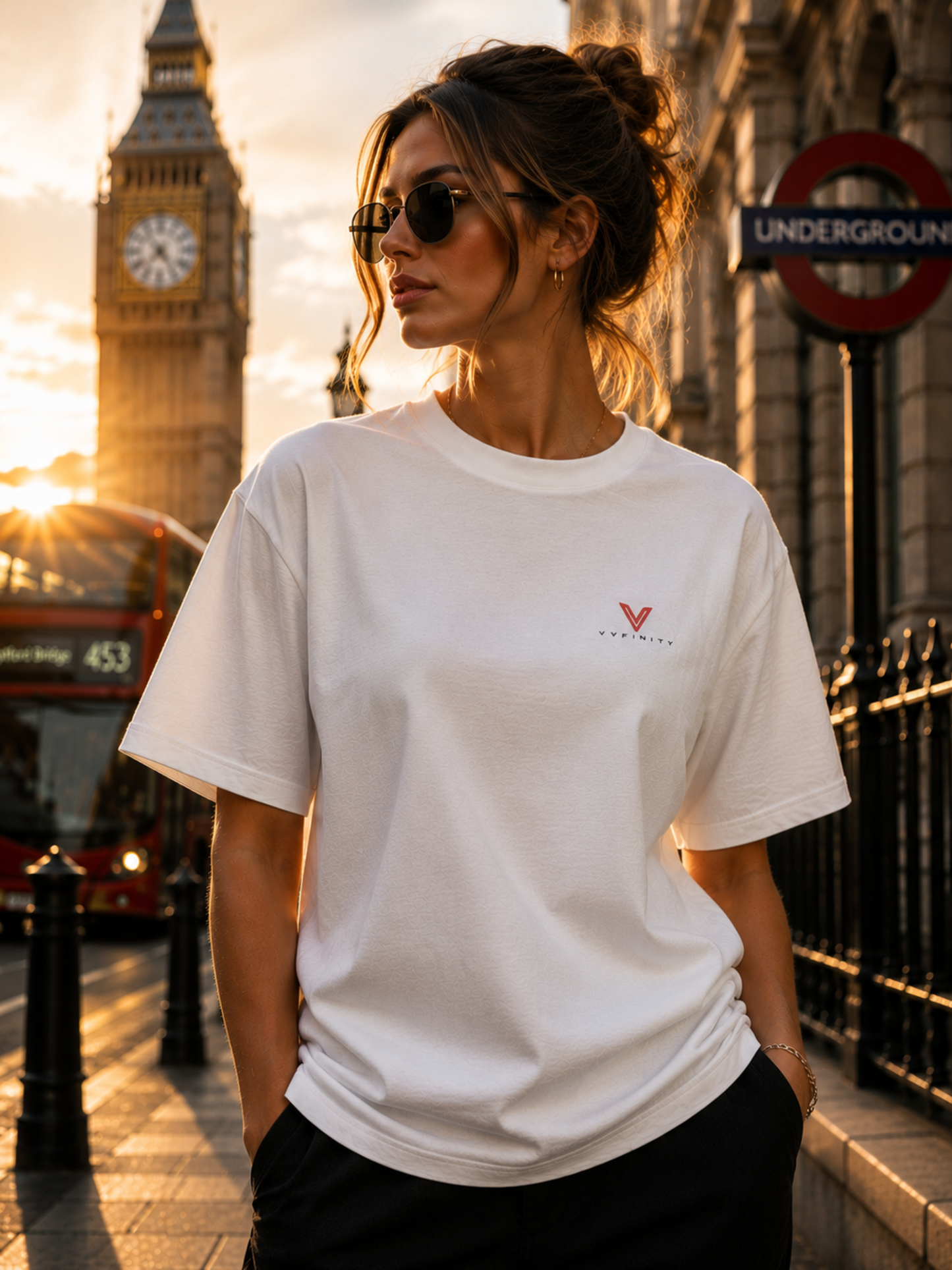 Frau trägt ein weißes VYFINITY Oversized Unisex T-Shirt mit rotem Brustlogo vor dem Big Ben und einer London Underground Station bei Sonnenuntergang. Modernes Streetwear Lifestyle-Foto mit rotem Doppeldeckerbus, urbanem City-Flair und minimalistischem Premium Fashion Look.