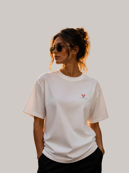 Frau trägt weißes VYFINITY Oversized Unisex T-Shirt mit kleinem rotem VYFINITY Logo auf der linken Brust. Minimalistisches Premium-Streetwear-Shirt mit lockerer Passform, kurzen Ärmeln und modernem Lifestyle-Look bei warmem Sonnenlicht.