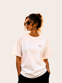 Frau trägt ein weißes VYFINITY T-Shirt mit kleinem rot-schwarzem Logo auf der Brust. Die minimalistische Frontansicht kombiniert einen lockeren Streetwear-Fit mit schwarzer Hose und Sonnenbrille vor einem warmen, neutralen Hintergrund.