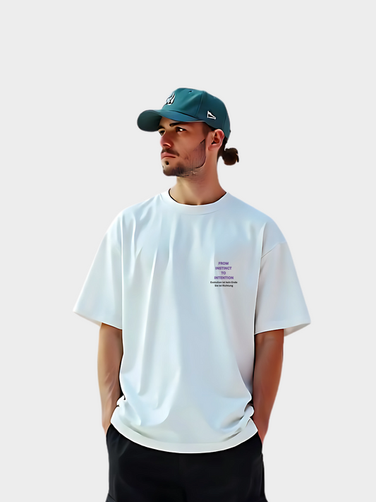 Oversized T-Shirt in Weiß mit kleinem Statement-Print „From Instinct to Intention“ auf der linken Brust. Getragen von einem Model mit dunkler Hose und grüner Cap. Lockerer Streetwear-Look für moderne Outfits und urbanen Lifestyle.