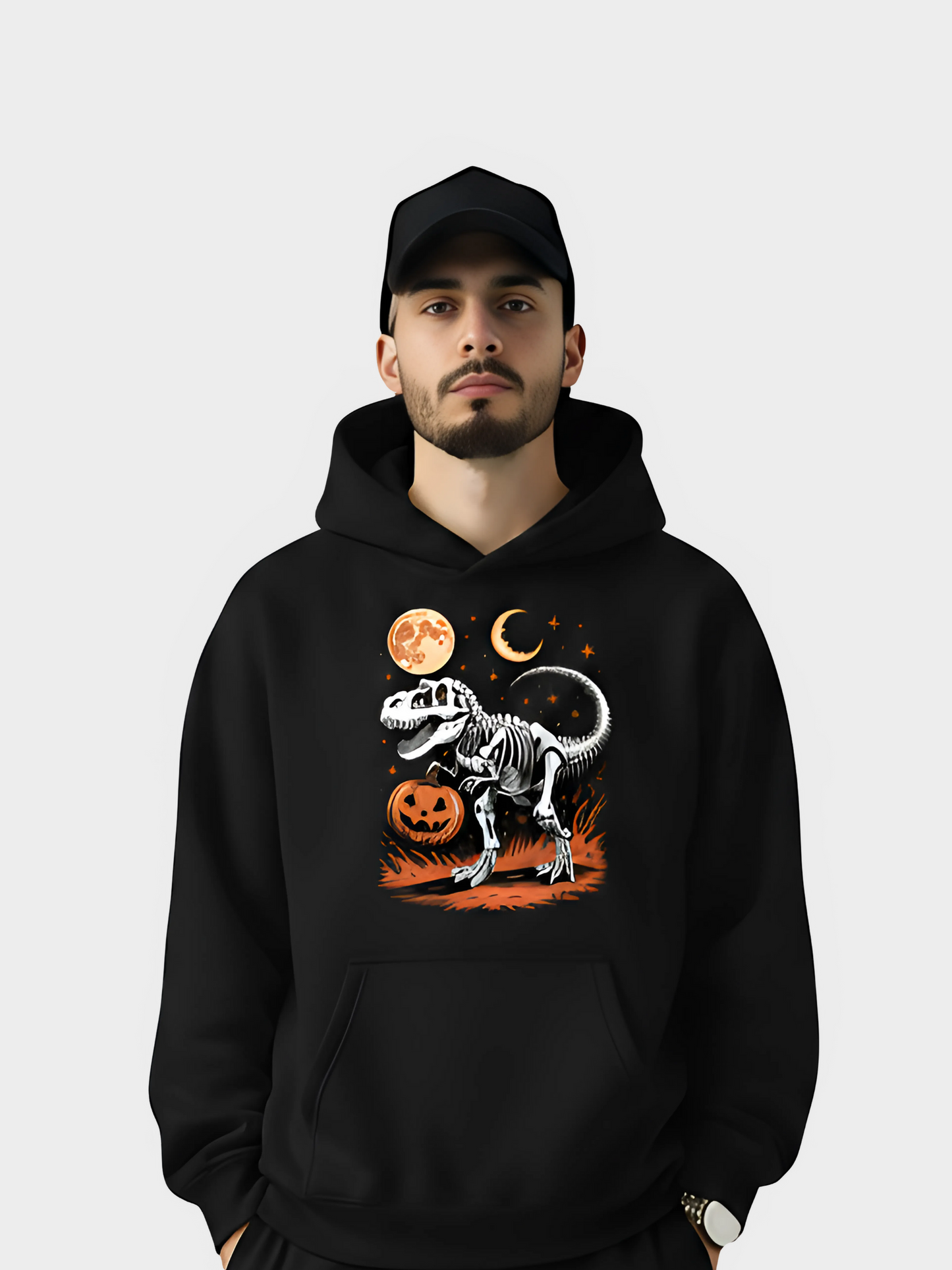 Schwarzer Hoodie mit Halloween-Motiv: T-Rex-Skelett vor Vollmond, Halbmond und Kürbis mit Gesicht. Getragen von einem Mann mit schwarzer Cap auf neutralem Hintergrund.
