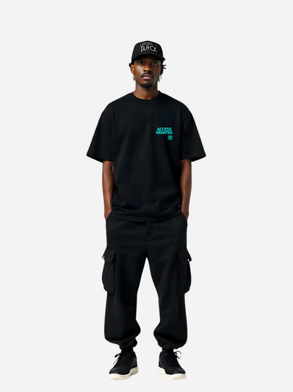 Model trägt ein schwarzes T-Shirt mit leuchtend cyanfarbenem Brustprint „ACCESS GRANTED“ und kleinem QR-Code. Streetwear-Stil mit lockerem Schnitt, kombiniert mit schwarzer Cargohose, Sneakers und Cap.