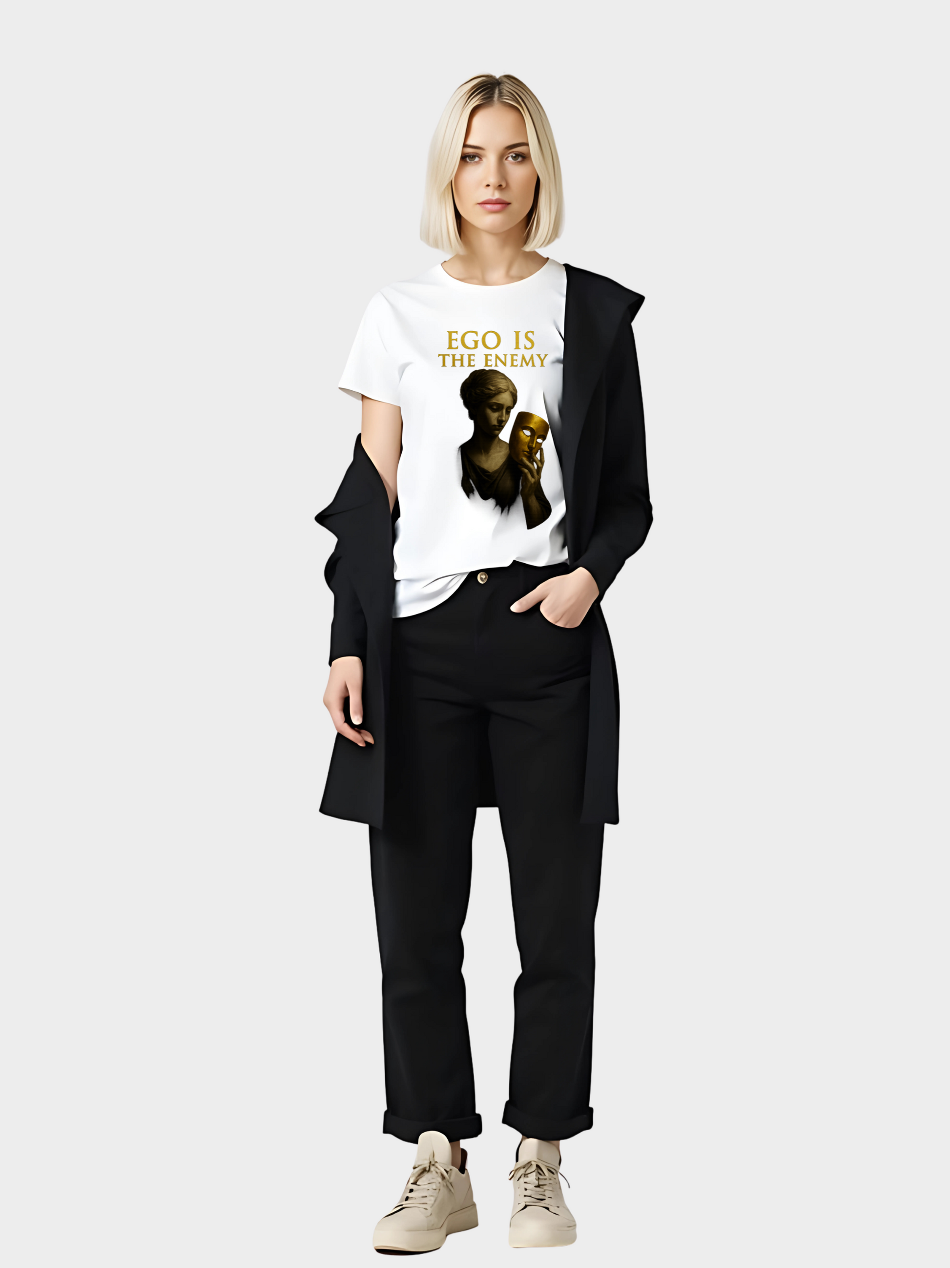 Moderne Frau trägt ein weißes T-Shirt mit goldenem Schriftzug „EGO IS THE ENEMY“. Design zeigt eine klassische Statue, die eine goldene Maske in der Hand hält. Gestylt mit schwarzer Hose, weiß-beigen Sneakern und schwarzem Blazer – eleganter, urbaner Look für Streetwear und Statement Fashion.