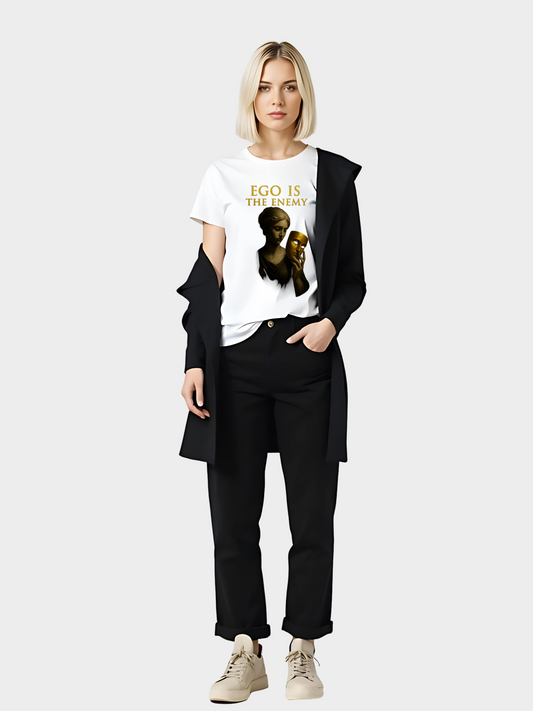 Moderne Frau trägt ein weißes T-Shirt mit goldenem Schriftzug „EGO IS THE ENEMY“. Design zeigt eine klassische Statue, die eine goldene Maske in der Hand hält. Gestylt mit schwarzer Hose, weiß-beigen Sneakern und schwarzem Blazer – eleganter, urbaner Look für Streetwear und Statement Fashion.