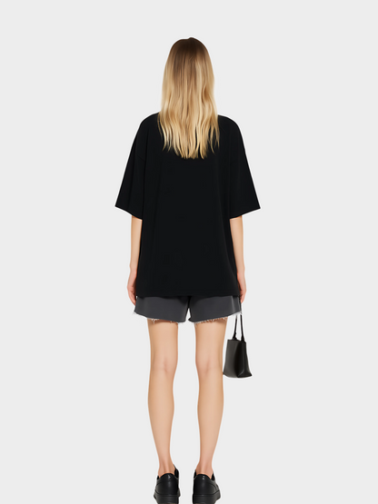 Model zeigt die Rückseite des schwarzen Oversize Vyfinity T-Shirts ohne Print. Lässiger Streetwear-Look mit grauen Shorts, schwarzen Sneakern und kleiner Handtasche, minimalistisch und modern.