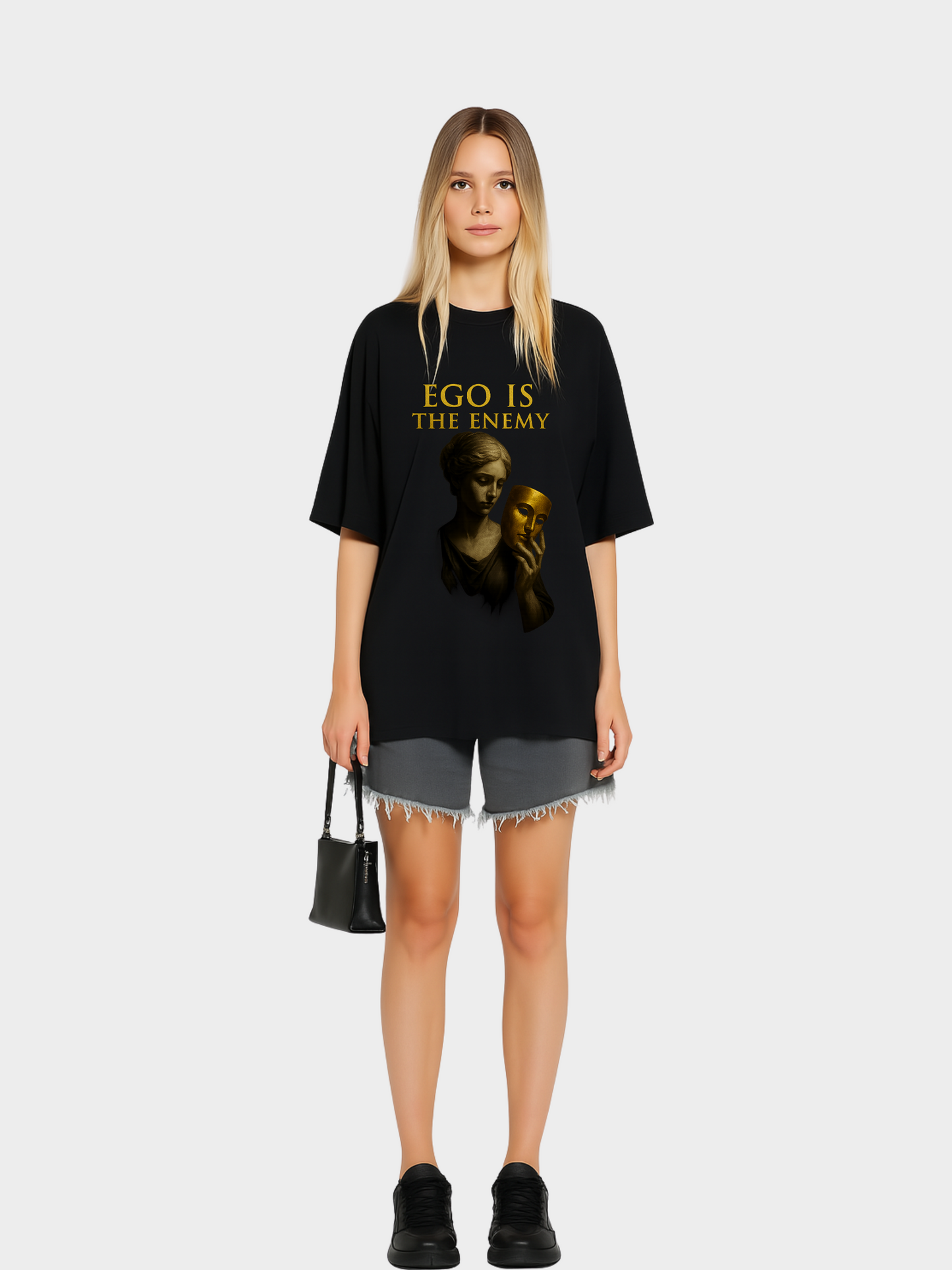 Model trägt schwarzes Oversize Vyfinity T-Shirt mit goldenem Aufdruck „EGO IS THE ENEMY“. Design mit antiker Statue, die eine goldene Maske in der Hand hält. Moderner Streetwear-Style, kombiniert mit grauen Shorts und schwarzer Handtasche.