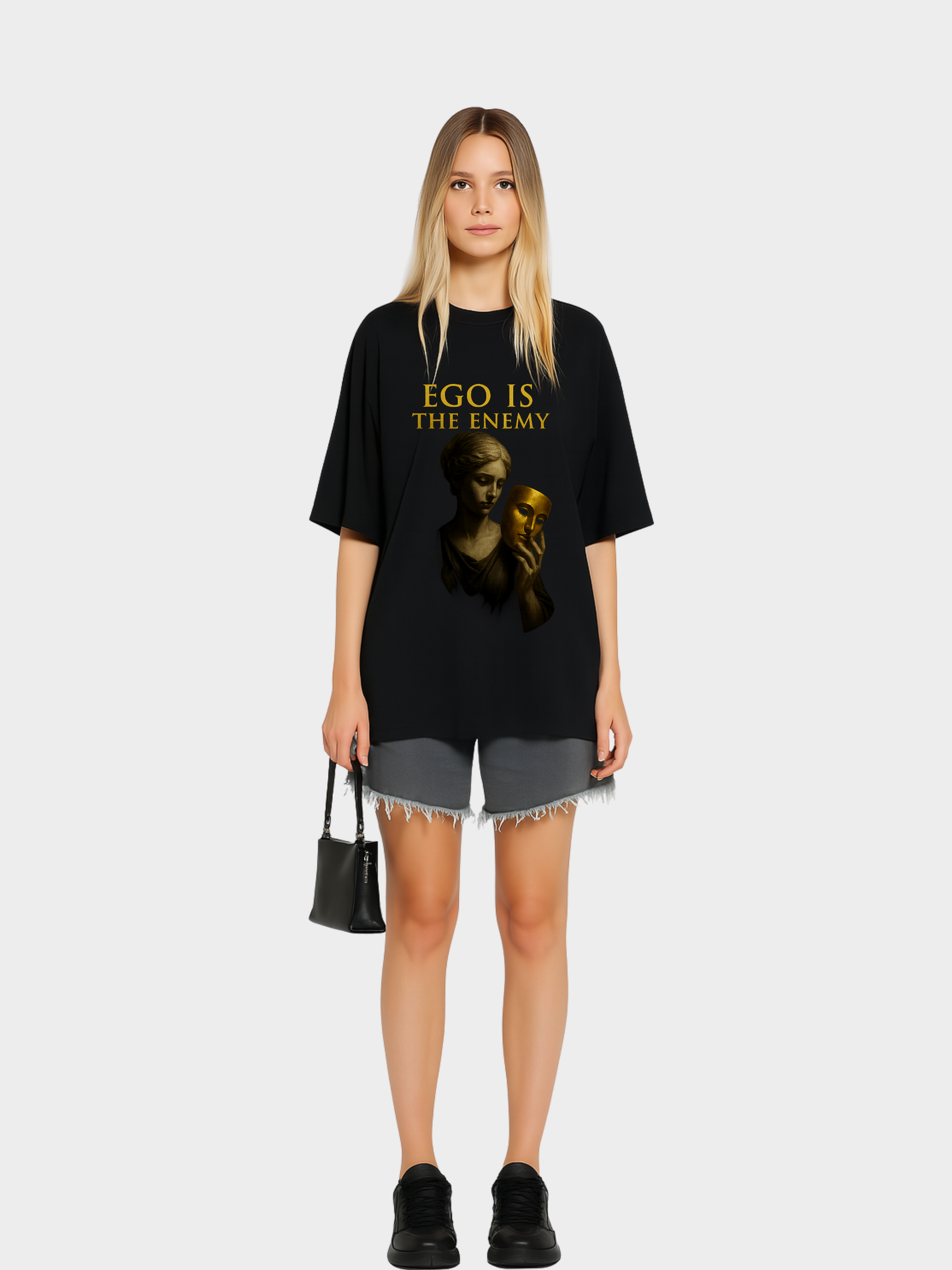 Model trägt schwarzes Oversize Vyfinity T-Shirt mit goldenem Aufdruck „EGO IS THE ENEMY“. Design mit antiker Statue, die eine goldene Maske in der Hand hält. Moderner Streetwear-Style, kombiniert mit grauen Shorts und schwarzer Handtasche.