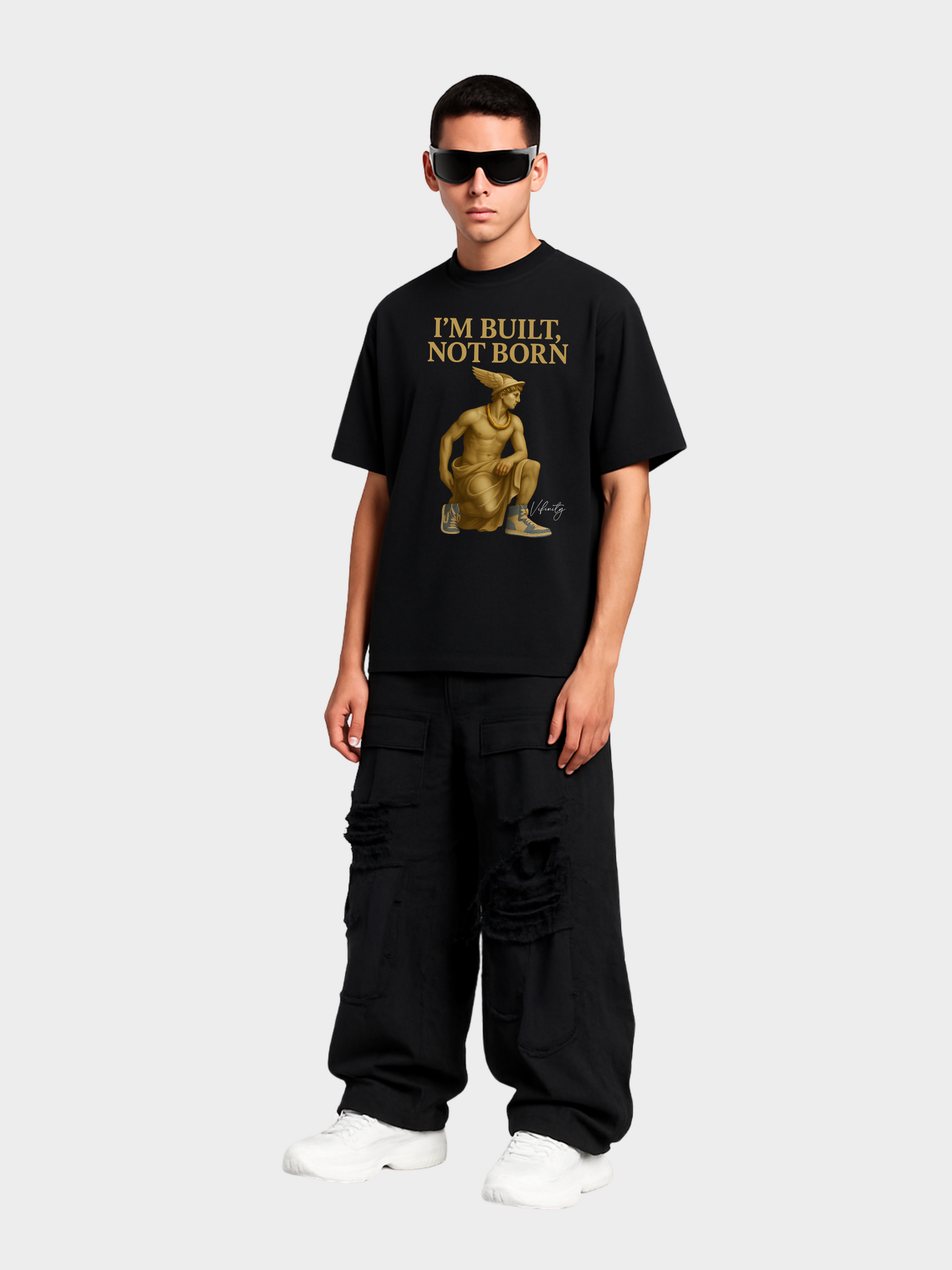 Schwarzes Oversize T-Shirt mit goldener Statue und Schriftzug „I’M BUILT, NOT BORN“. Getragen von männlichem Model mit schwarzen Cargo Pants und weißen Sneakern. Moderner Streetwear-Look mit urbanem Style.