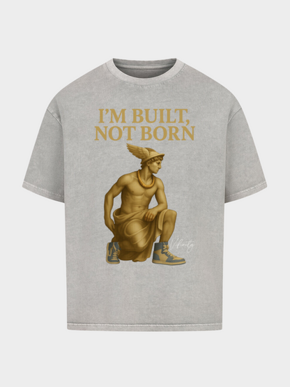 Graues Oversize T-Shirt im Washed-Look mit goldener Statue im antiken Design, kombiniert mit modernen Sneakers. Oben der goldene Motivationsschriftzug „I’M BUILT, NOT BORN“ – stilvolle Streetwear mit antikem Flair.