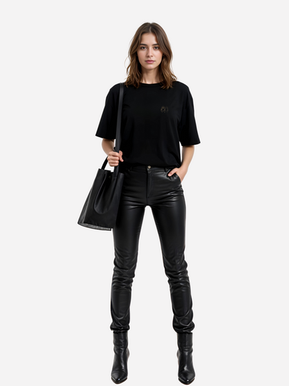 Vyfinity Oversize T-Shirt in Schwarz mit kleinem Line-Art Gesicht auf der Vorderseite, getragen von Frau mit Lederhose und schwarzer Tasche. Modernes Streetwear Outfit für urbanen Lifestyle.